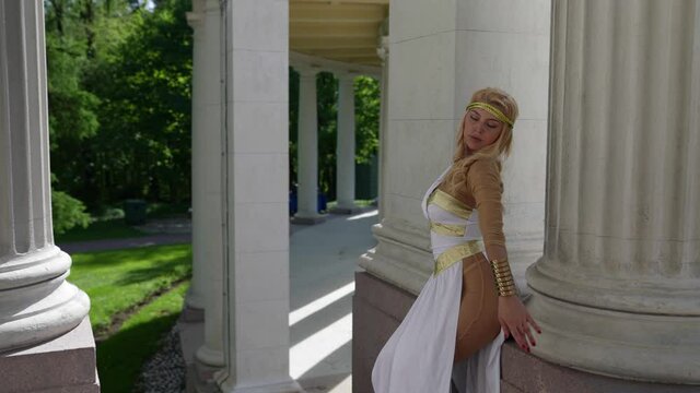 sexy blonde woman is wearing suit of Aphrodite is posing sexually near columns of palace