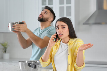 Young man collecting leaking water from ceiling while his girlfriend calling roof repair service in kitchen