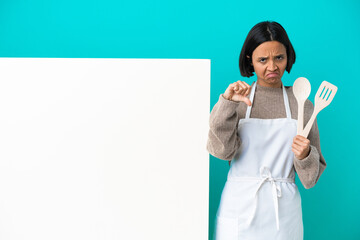 Young mixed race cook woman with a big placard isolated on blue background showing thumb down with...