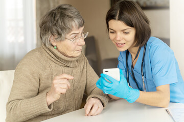 Obraz premium A nurse in a medical suit takes care and explains to an elderly patient how to use applications on a smartphone. Grandmother 80+ years old, does not understand how to communicate with a doctor online.