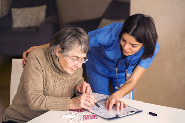 An elderly gray-haired woman with glasses reads a health insurance contract and signs it. The document is offered by a young woman to a doctor, a nurse with a stethoscope. Safety, care and medicine.