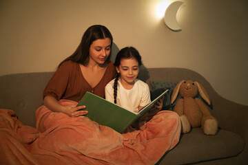 Little girl with mother reading book in living room lit by night lamp