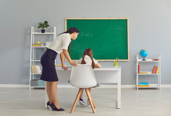 Young woman teacher standing and checking girl pupil sitting at desk and doing homework in school with school furniture at background, rear view. Education, children classes and school concept