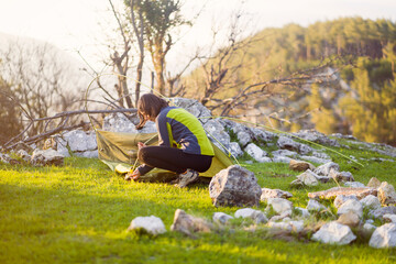 A young girl sets up her tent at a campsite on a green meadow among the mountains on a warm summer evening. The traveler is getting ready for a comfortable sleep in nature.