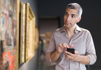 Adult man is visiting museum with mobile and looking at the pictures in the gallery indoor.