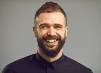 Close up portrait of a bearded man in a black shirt who is sincerely smiling and has wrinkles around his eyes. Concept of positive human emotions and people. Gray background. Banner. Place for text.