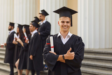 Young smiling student in a black robe and academic hat stands with a diploma in his hands on the university stairs on graduation day. Guy looks into the camera and raises his thumb up.