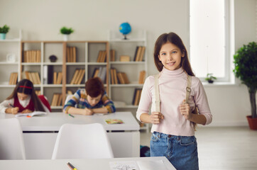 Fototapeta na wymiar Happy smiling caucasian girl pupil stand with backpack next to classroom desk. Portrait of elementary school student looking at camera with classmate on blurred background. Back to school, education