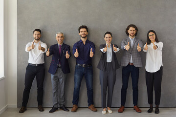 Happy business leaders or group of employees showing thumbs up while standing against gray wall and...