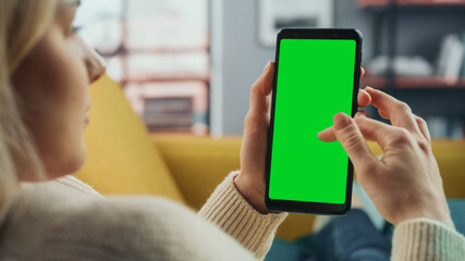 Beautiful Authentic Female Using a Smartphone with Green Screen Mock Up Display at Home Living Room...