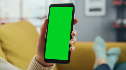 Beautiful Authentic Female Using a Smartphone with Green Screen Mock Up Display at Home Living Room while Lying on a Couch Sofa. She's Browsing the Internet and Checking Videos on Social Networks.
