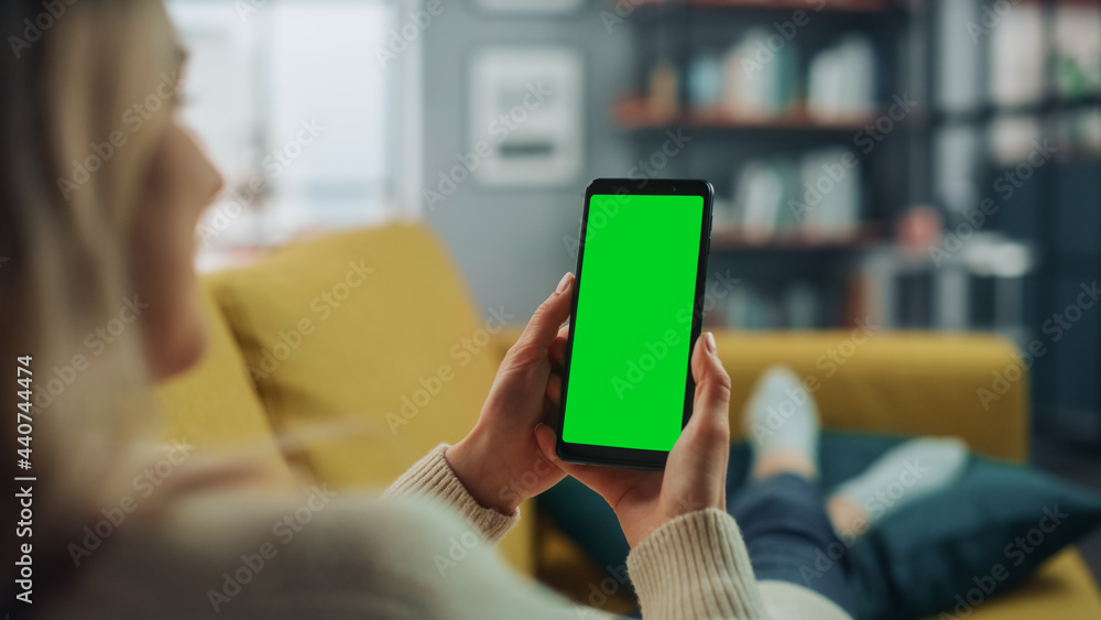Aufkleber Beautiful Authentic Female Using a Smartphone with Green Screen Mock Up Display at Home Living Room while Lying on a Couch Sofa. She's Browsing the Internet and Checking Videos on Social Networks.
