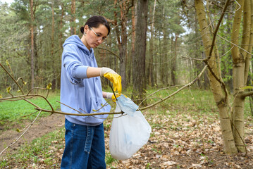 Woman in casual wear, gloves, with plastic trash bag collecting garbage in forest. Plogging