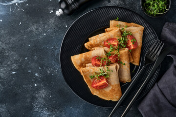 Stack of gluten free buckwheat flour crepes pancakes with cherry tomatoes and arugula microgreens on black plate on dark background, homemade healthy baking for breakfast. Mock up. Copy space.
