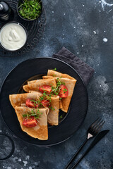 Stack of gluten free buckwheat flour crepes pancakes with cherry tomatoes and arugula microgreens on black plate on dark background, homemade healthy baking for breakfast. Mock up. Copy space.