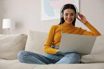 Woman with laptop and headphones sitting on sofa at home - obrazy, fototapety, plakaty