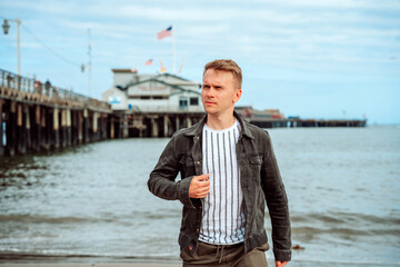 A stylish young man in a denim jacket walks along the beach against the backdrop of the pier and the ocean in Santa Barbara, California, USA