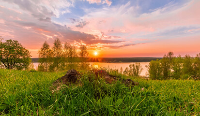 Fototapeta na wymiar Scenic view at beautiful sunset on a shiny lake with old rough stone on the foreground, green grass, birch trees, golden sun rays, calm water ,nice cloudy sky on a background, spring landscape