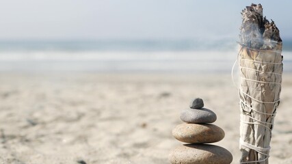 Dried white sage smoke, smudge stick burning in soft focus with bokeh, aroma smudging close up. Rock balancing on ocean beach, stones stacking by sea water waves. Zen pyramid of pebbles on sandy shore