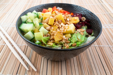 Vegetarian poke bowl in design black bowl with chopsticks below, on a reed pad background. Green cucumber, red pepper, peanuts, corn, beetroot on top of sushi rice. Healthy vegetarian food.