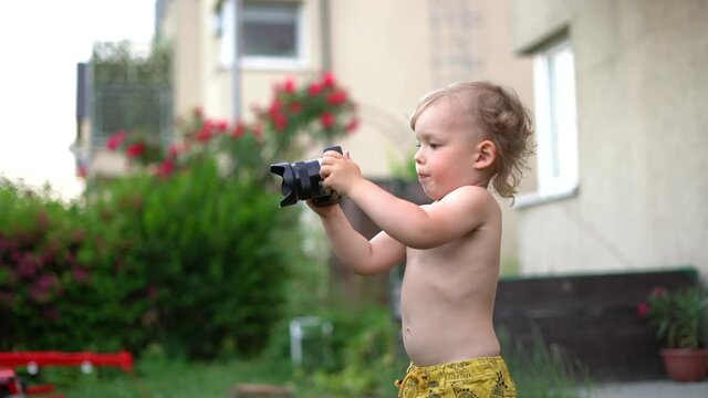 Three-year-old is learning to photograph. The child holds a professional camera in his hands and aims the lens at objects around in the yard. Summer vacation. Education concept