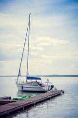 Lonely small sailing white yacht is moored at a wooden pier on the lake on a summer day. Vertical photo