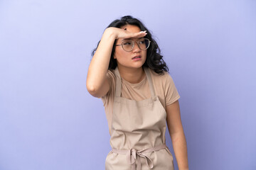 Restaurant asian waiter isolated on purple background looking far away with hand to look something