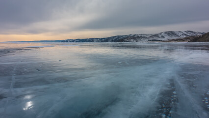 Obraz premium Sunset over a winter lake. The cloudy sky is highlighted in orange. Cracks on the smooth ice, bubbles of frozen methane gas. Reflections of the sun on the surface. Mountains on the horizon. Baikal