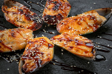 Shellfish mussels Baked with cheese in a shell on a black stone. Grilled mussels with teriyaki sauce and sesame seeds. Seafood lunch, close-up