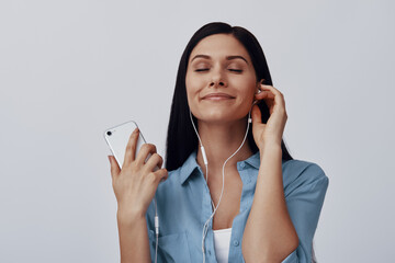 Attractive young woman listening music and smiling while standing against grey background