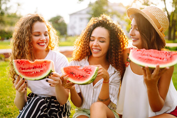 Three young woman  in the park sitting on the grass in joyful in sunny day and eating watermelon. Female friends relaxing and enjoying holidays together. People, lifestyle, travel, nature and vacation