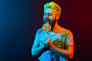 Handsome young adult bearded hipster man with lolly pop in hands, enjoying licking tasty candy,...