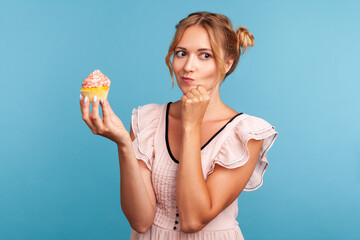Pensive young adult blonde female with funny hair buns holding cake in hand and thinks eat or not,...