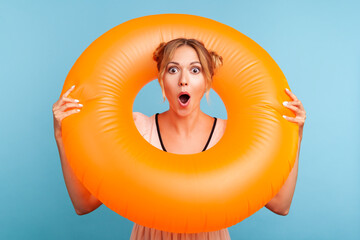 Shocked female with blonde hair and two hair buns holding rubber ring over her neck and looking at camera with surprised expression and opened mouth. Indoor studio shot isolated on blue background.