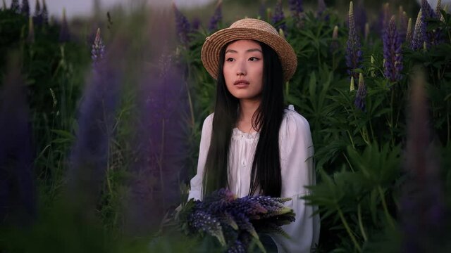 Beautiful asian woman in straw hat in the flowers field on sunset. Young girl sitting on a flower glade outdoors