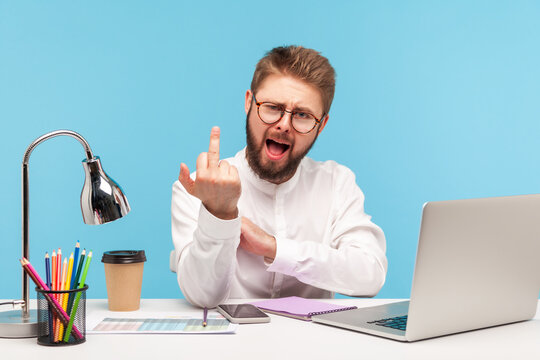 Excited rude man with beard office worker showing middle finger looking at camera sitting at workplace, uncultured gesture, disrespect and negativity. Indoor studio shot isolated on blue background