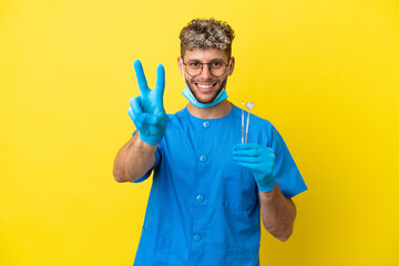 Dentist caucasian man holding tools isolated on yellow background smiling and showing victory sign