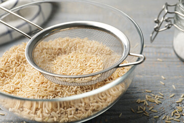 Sieve with rice in bowl on grey wooden table, closeup