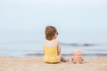 Little girl in yellow swimsuit and brown teddy bear sitting on beach sand and staring at sea water...