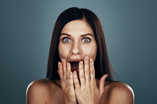 Surprised Young Woman Looking At Camera And Covering Mouth With Hand While Standing Against Grey Background