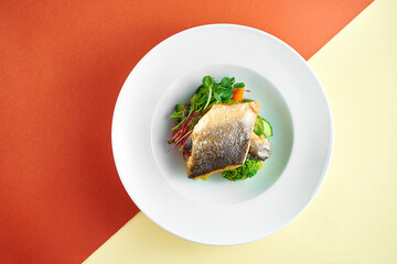Fried sea bass fillet with steamed vegetables for garnish in a white plate on colored backgrounds.