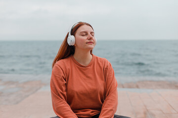 Beautiful caucasian young woman meditates with wireless headphones on the beach near the sea