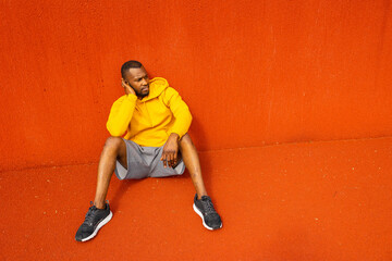 Tired pensive athlete sportsman sitting isolated near red wall, having rest after workout,...