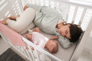 Obraz na płótnie Canvas Tired young mother sleeping in crib with her baby at home, above view