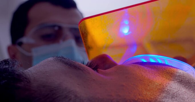 A close-up male patient with optragate on a mouth is having treatment by using curing light for solidification filling on a tooth. In the background is a male medical worker in a mask.