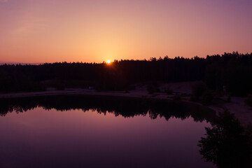 The sun shines from behind the silhouettes of the pine forest, which is reflected in the mirror water of the lake