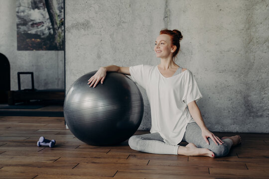 Happy fitness woman elegantly sits on parguet floor leaning one hand on silver fitball