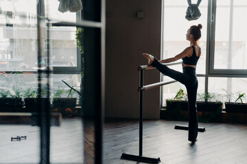 Slim ballerina during her stretch routine next on ballet barre in studio - obrazy, fototapety, plakaty
