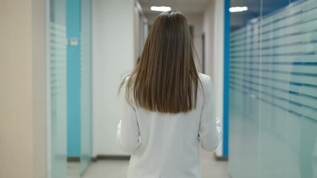 Back view of slim brunette young woman walking in office hallway with colleagues passing. Live camera follows confident Caucasian employee strolling in corridor at workplace. Business lifestyle