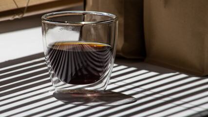 Coffee. An Americano in a glass beaker stands against the backdrop of a shuttered window and craft packaging.
